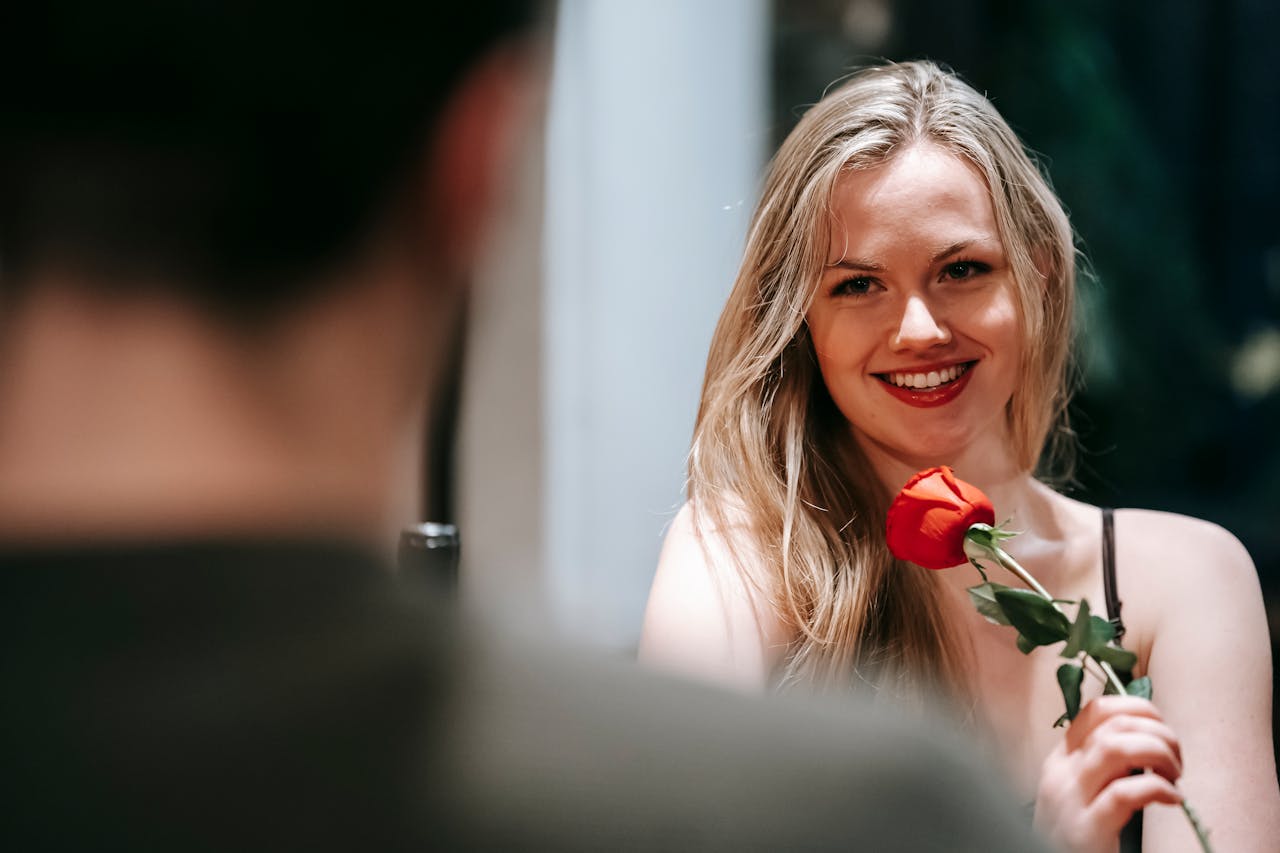 Young couple in elegant clothes sitting in light restaurant while enjoying date with red rose and looking at each other